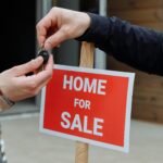 Two people exchange keys in front of a “Home for Sale” sign