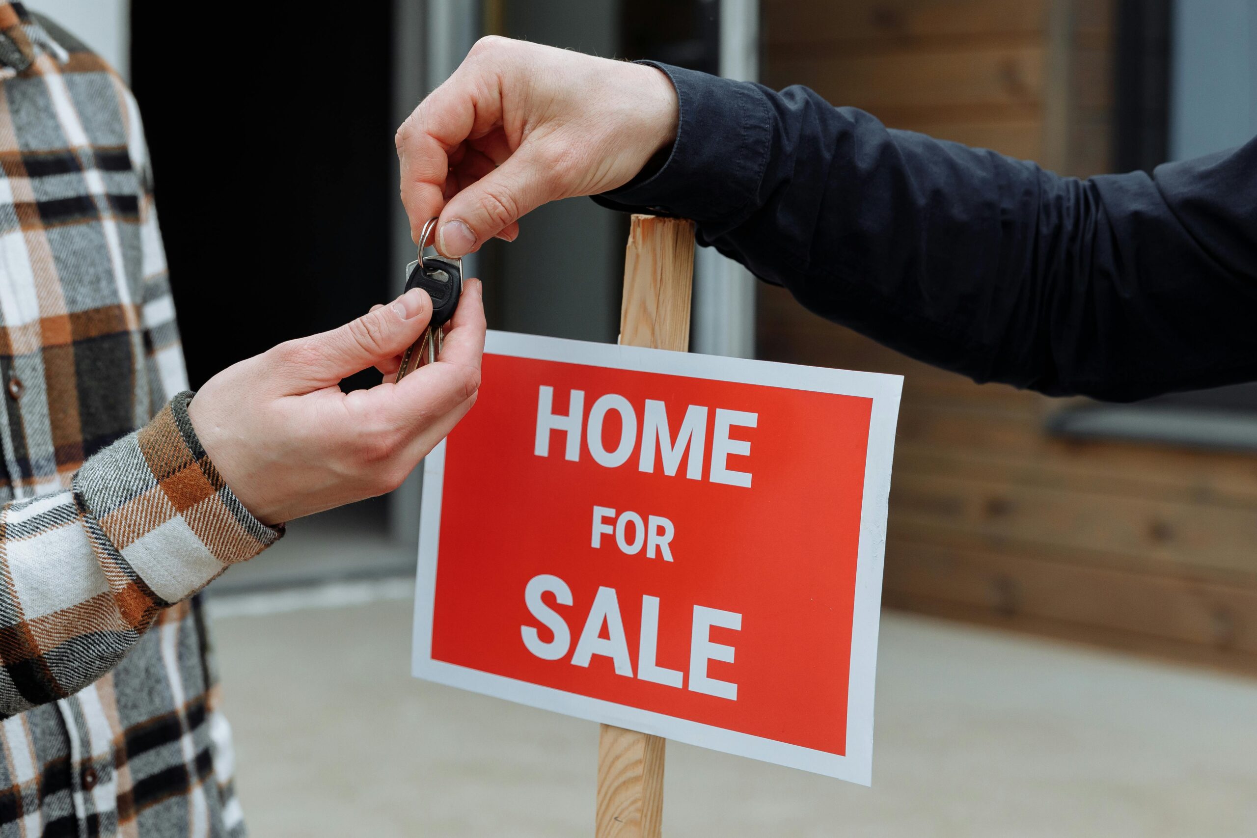 Two people exchange keys in front of a βHome for Saleβ sign