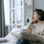 A woman in a white sweater gazes into the distance from a sofa