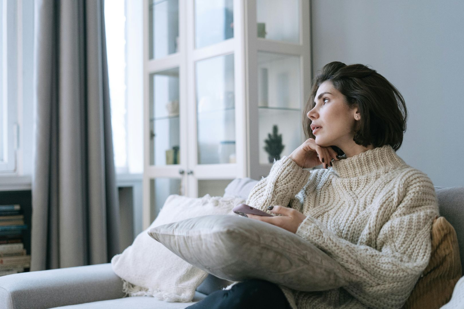 A woman in a white sweater gazes into the distance from a sofa