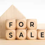 Plain wooden letter blocks that spell out “FOR SALE” in front of an unadorned wooden house figure