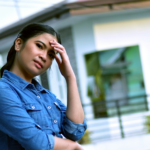 A woman stands in front of her house with her hand on her head