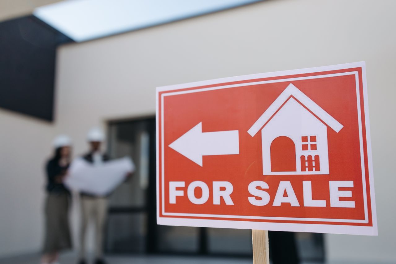 A for sale sign pointing to a house with two people standing in front
