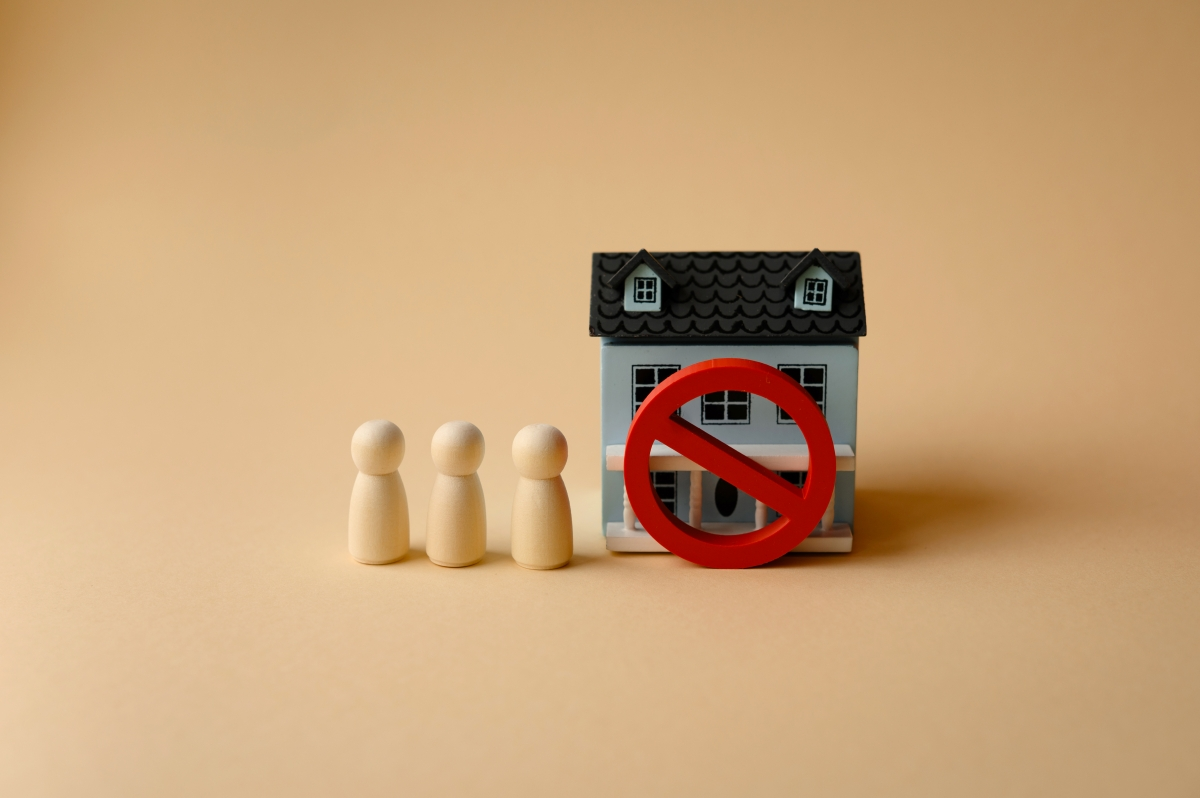 A house figure with a red prohibition sign in front of it and three wooden figures to the left