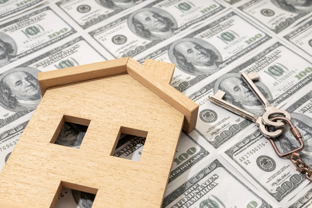 A flat wooden house figure and keys atop a background of continuous $100 bills