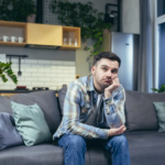 A pensive man sits on the sofa, resting his face on his hand