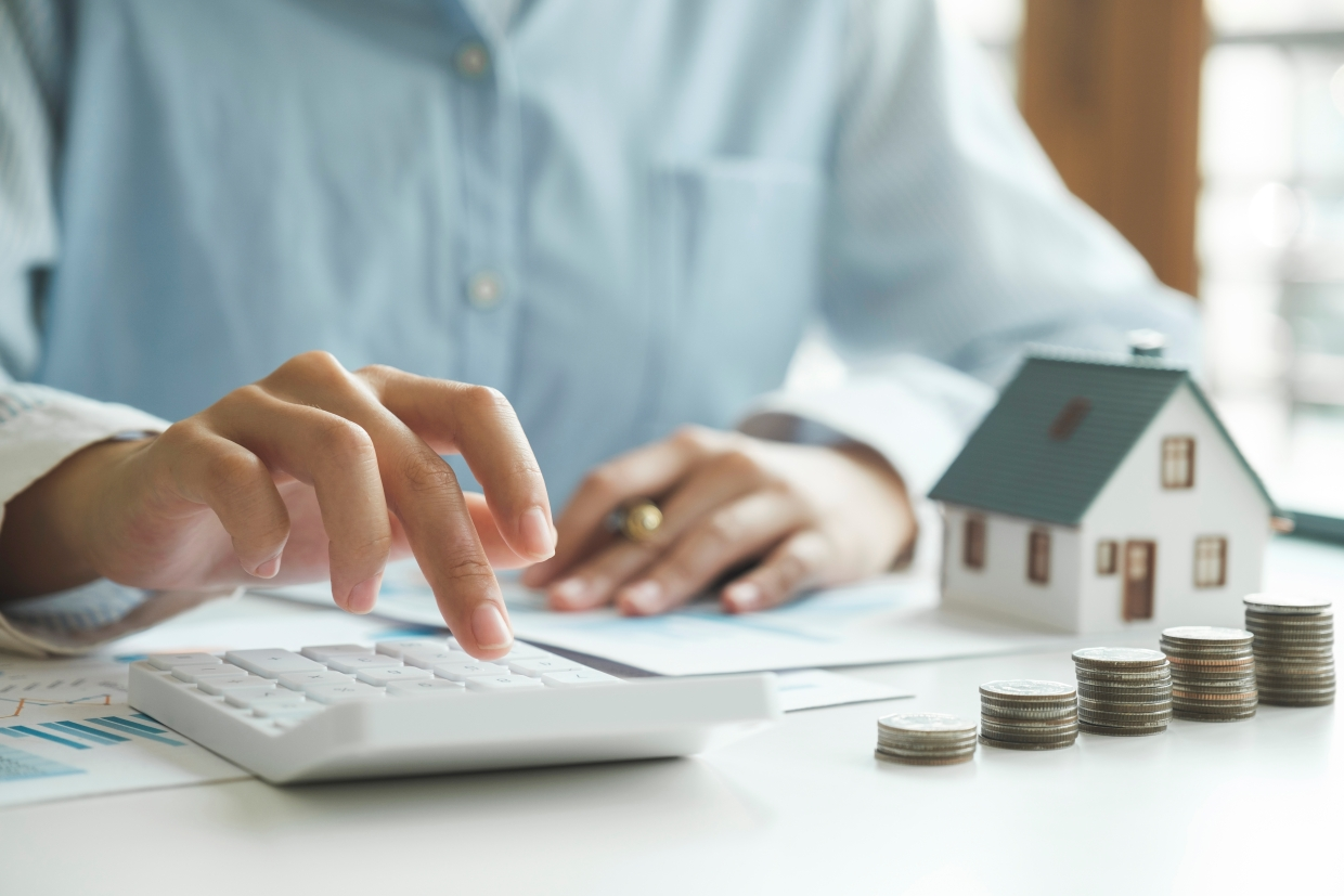 A seller uses a calculator to figure their costs, with a small house model and stacks of coins nearby