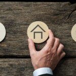 A hand holds a round token with a house illustration between two other tokens representing “BUY” and “SELL”