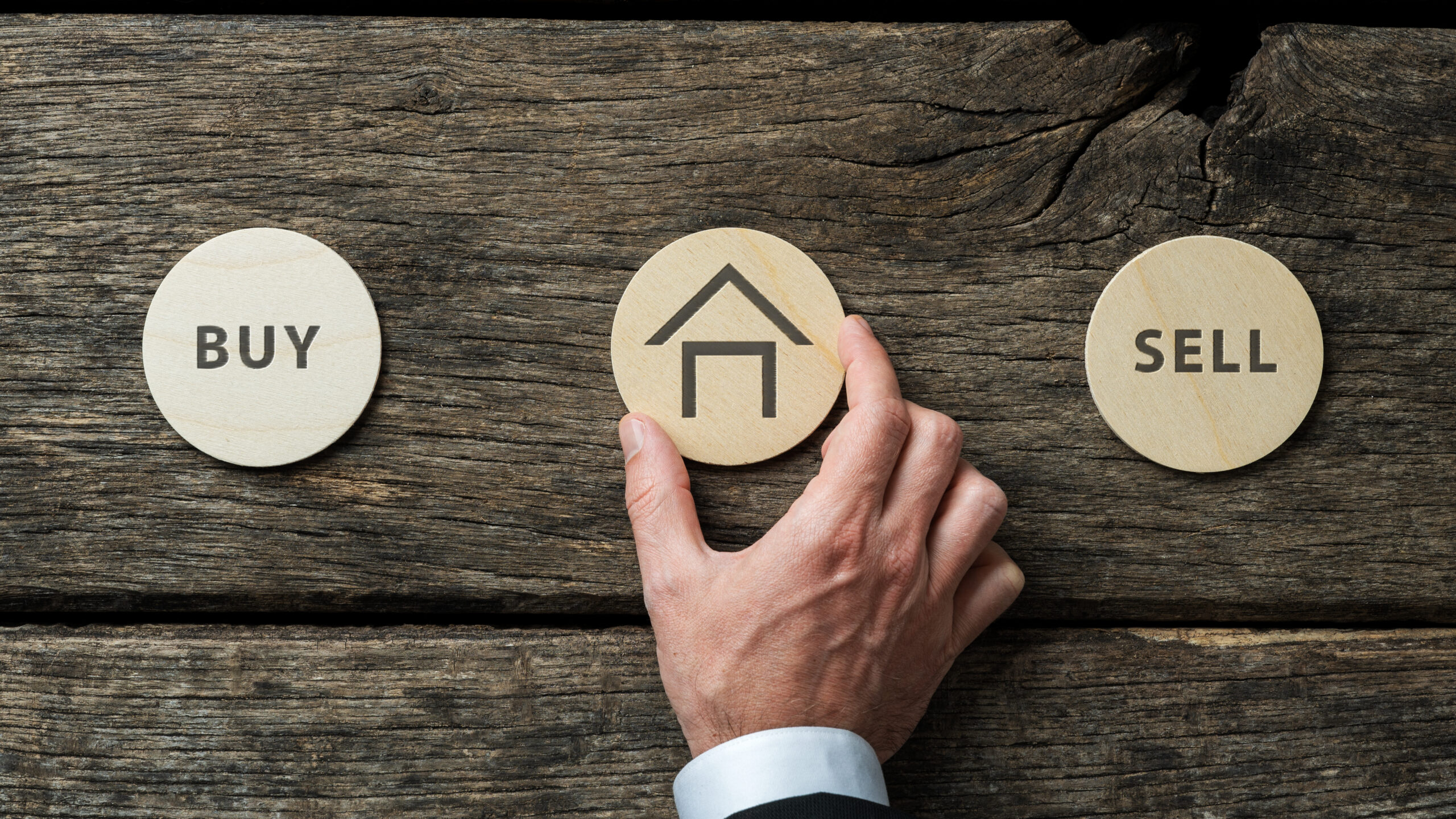 A hand holds a round token with a house illustration between two other tokens representing “BUY” and “SELL”